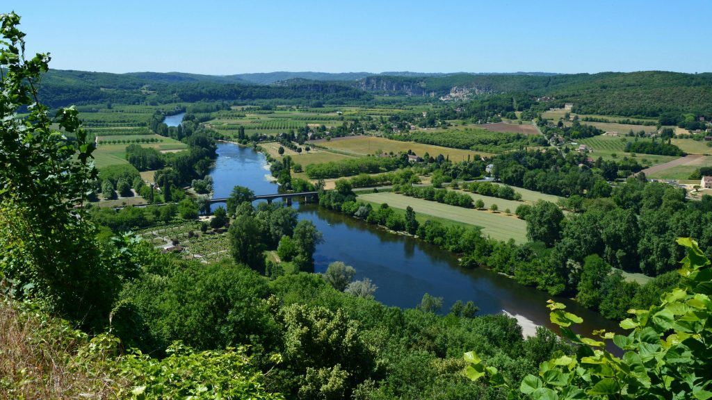 Aerial view of the Dordogne River surrounded by lush greenery and farmlands under a clear blue sky.