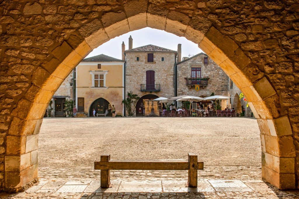 A picturesque view of a medieval square in Monpazier, France captured through a stone archway.