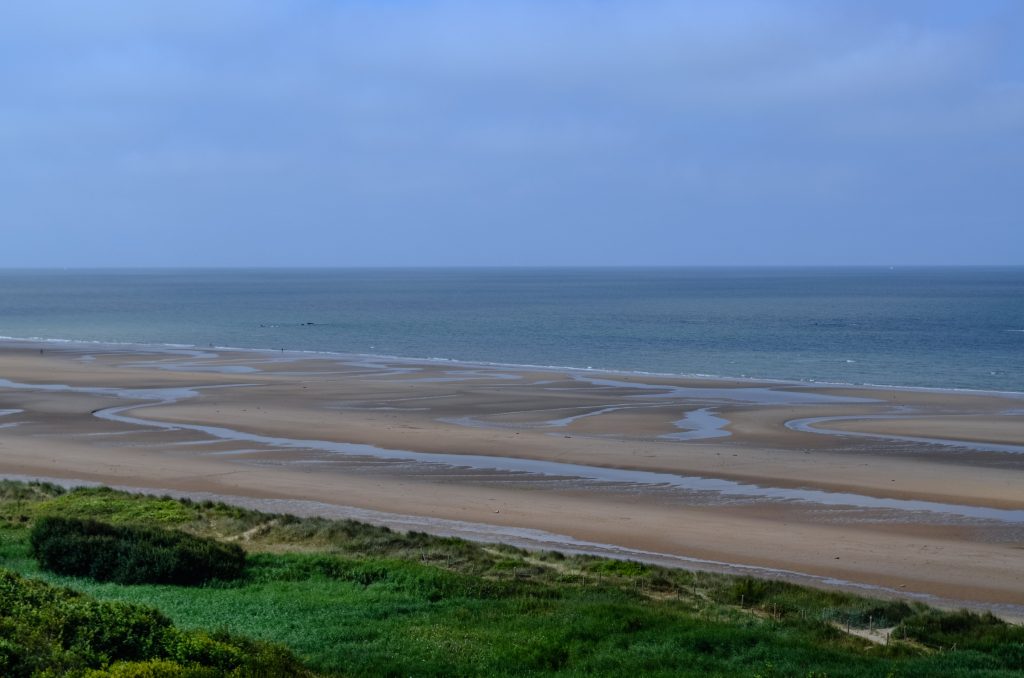 Tranquil view of Omaha Beach in France, showcasing vast sand and calm sea under blue skies.